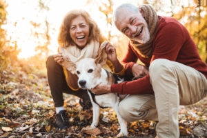 An elderly couple plays with their old dog among some autumnal trees. With pet insurance, they can enjoy their retirement funds while protecting their old dog from various health issues like joint problems, heart problems or cancer.